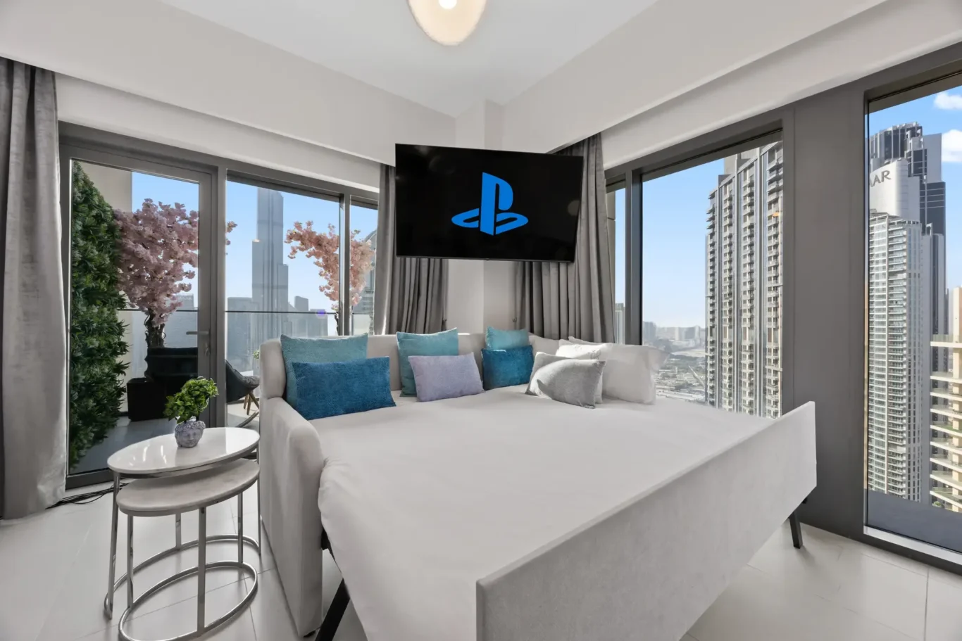 Bright hotel living area with a white sofa and blue cushions, a wall-mounted TV showing the blue PlayStation logo, and large windows overlooking city skyscrapers.