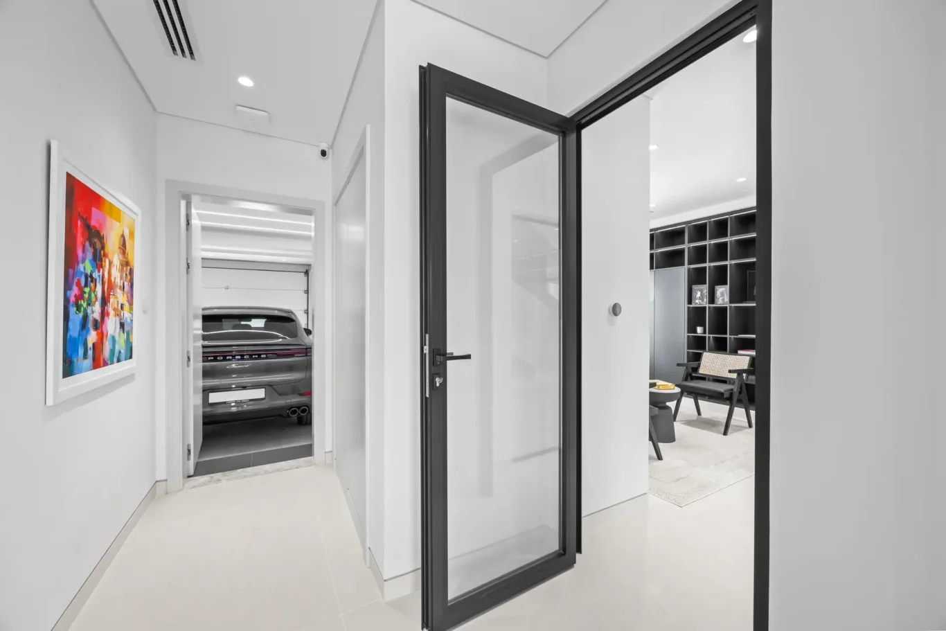 Open glass door with a black frame leading from a bright hallway to a room with dark shelving; colorful abstract painting on the left wall remains visible in grayscale.