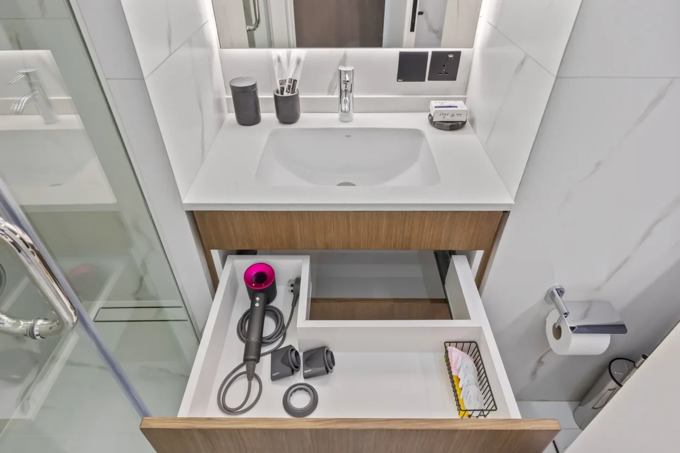 Modern bathroom vanity with white countertop; two open drawers reveal a Dyson hair dryer and attachments, toothbrush holder, and soap tray on the counter.