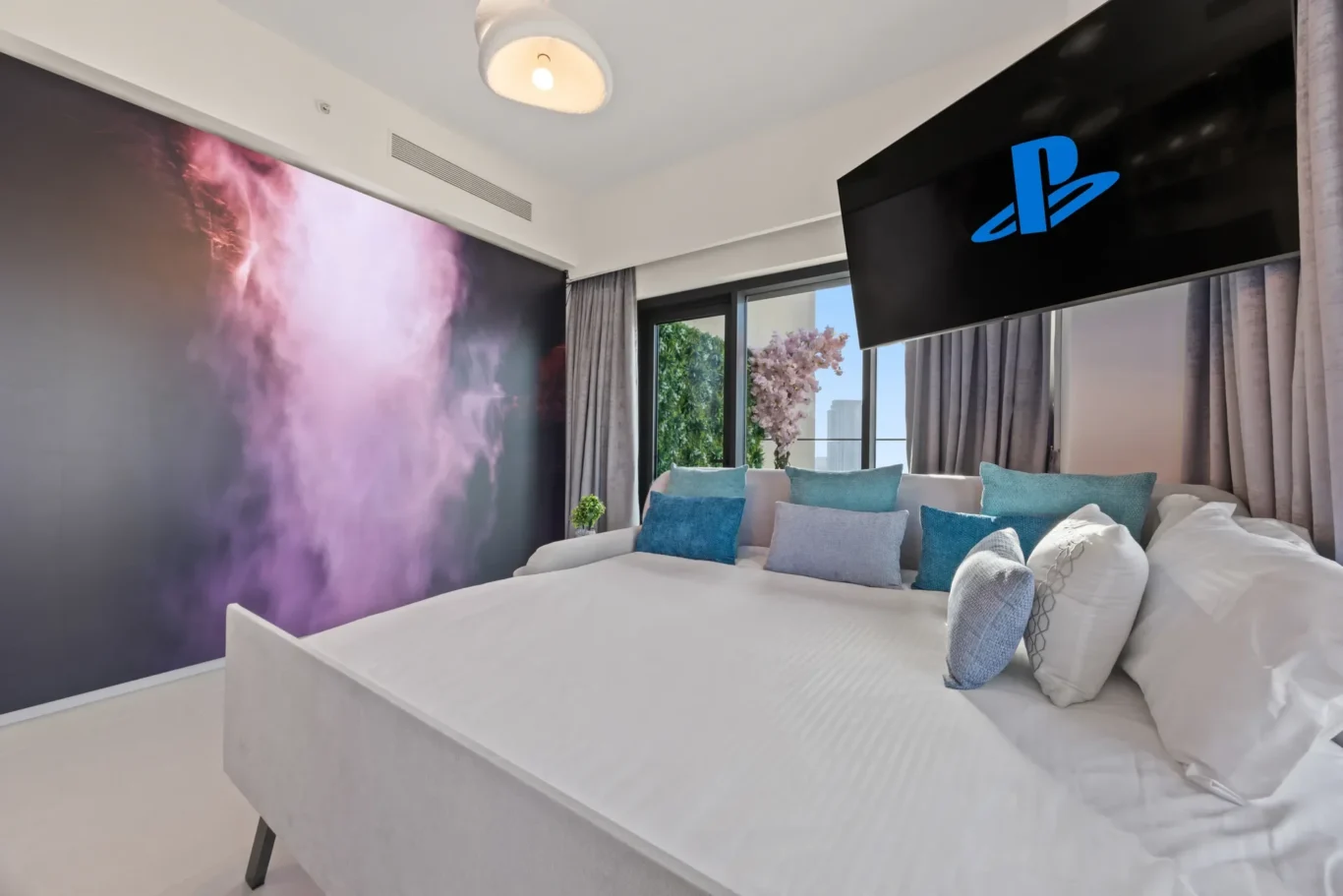 White bed with blue pillows in a modern room, a purple smoke mural on the wall, and a large wall-mounted TV displaying the PlayStation logo.
