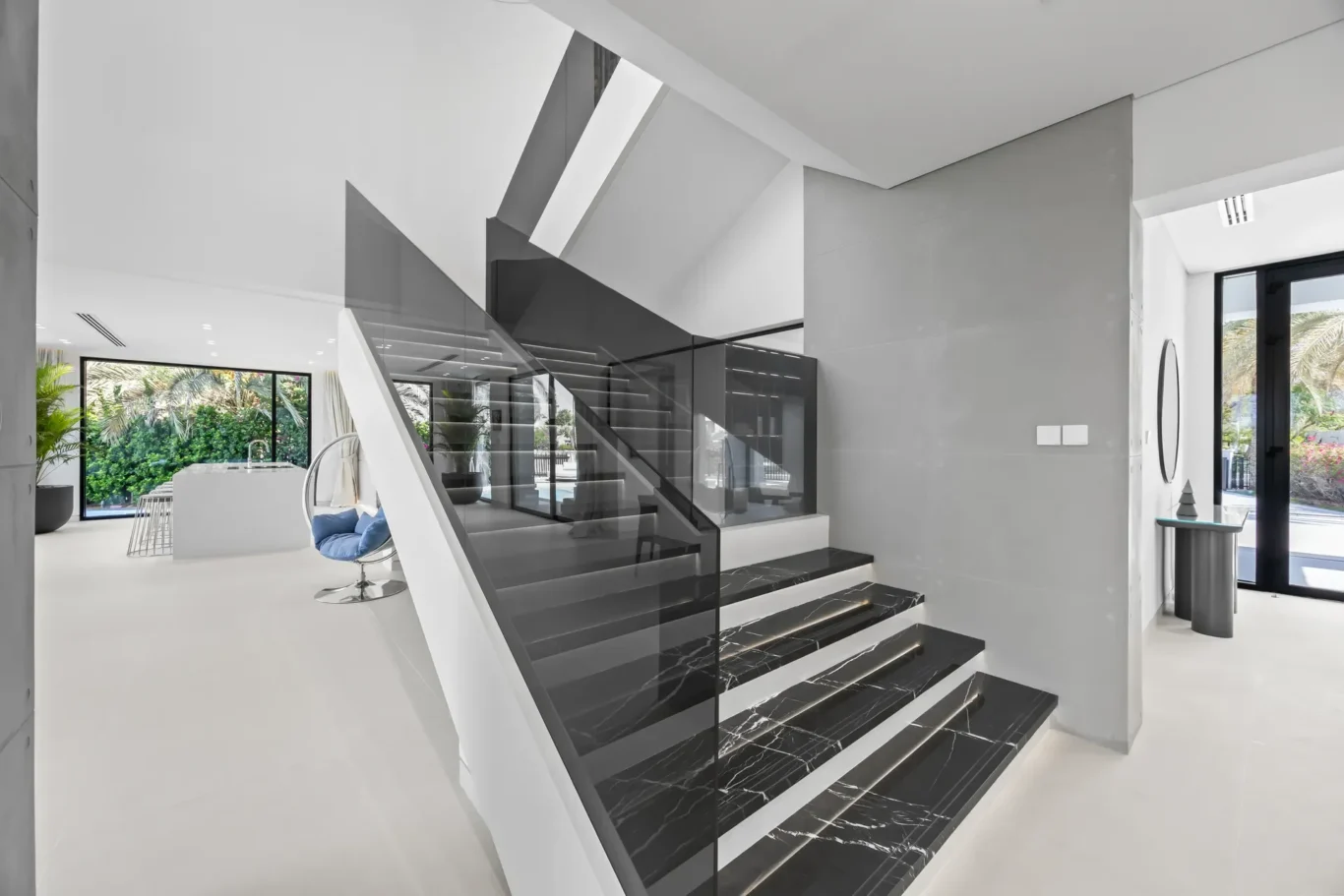 Modern open-concept foyer with black marble staircase and glass railing, white walls, and large windows overlooking greenery outside the home interior.