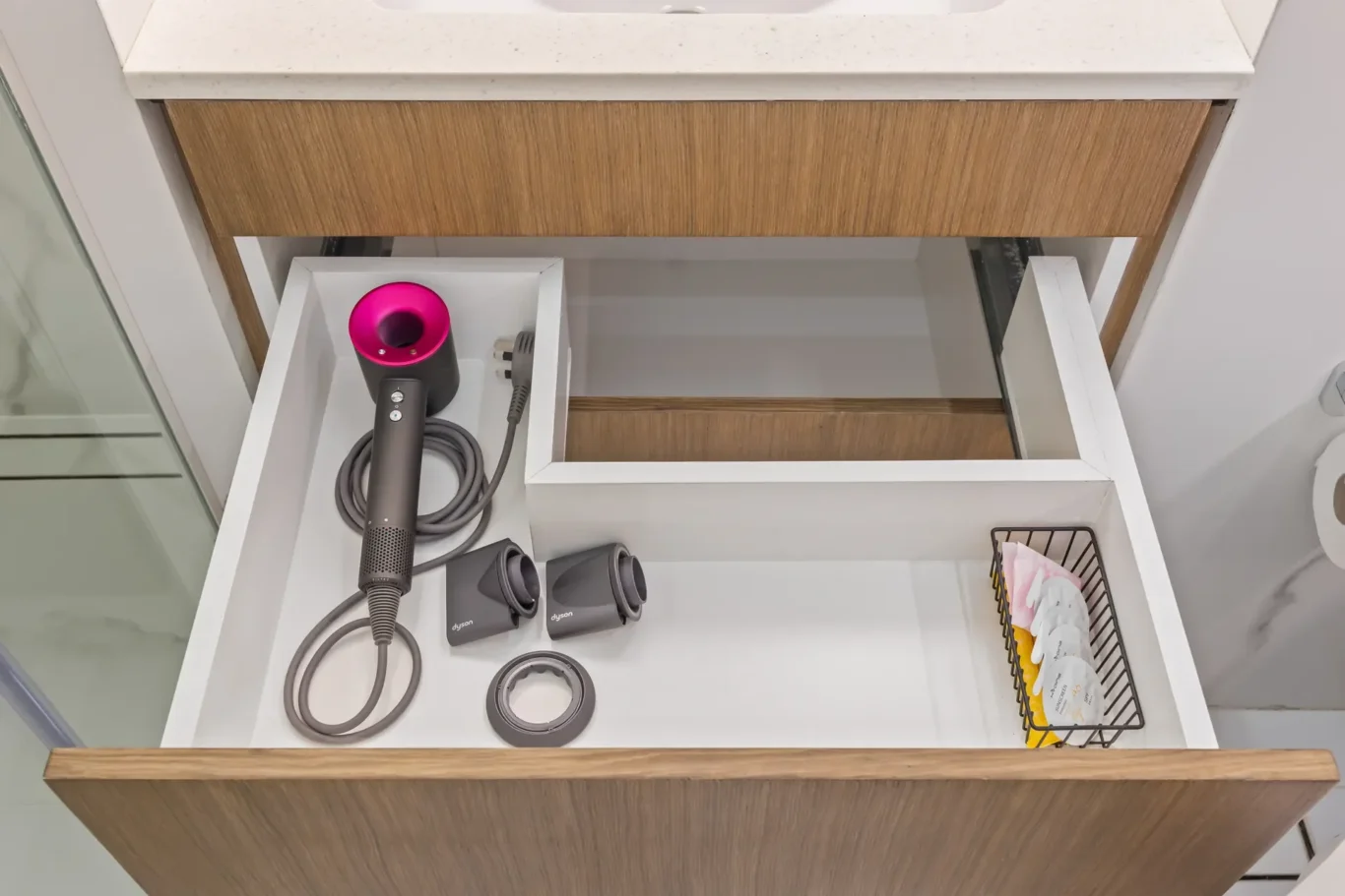 Drawer opened to reveal a Dyson hair dryer with pink nozzle and multiple attachments in a white tray under a wooden vanity top.