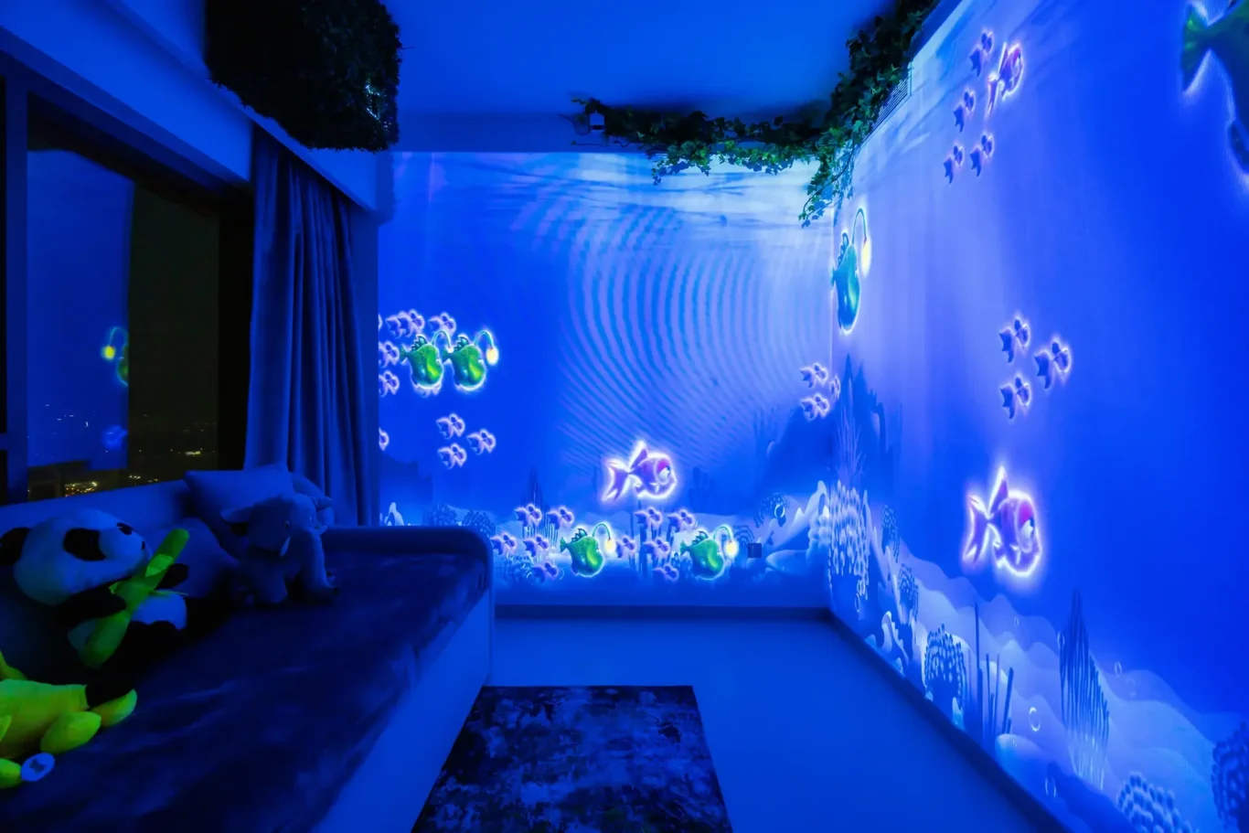 Blue-lit kids' bedroom with an underwater wall projection of fish and plants, plus stuffed animals on a sofa beside a window.
