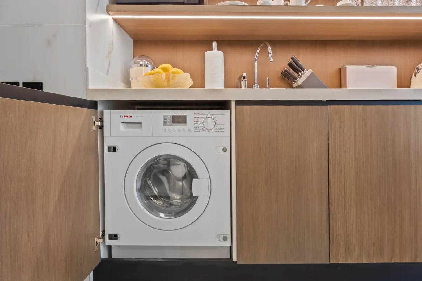 Washing machine built into kitchen cabinetry under a white countertop with wooden cabinet doors on both sides.