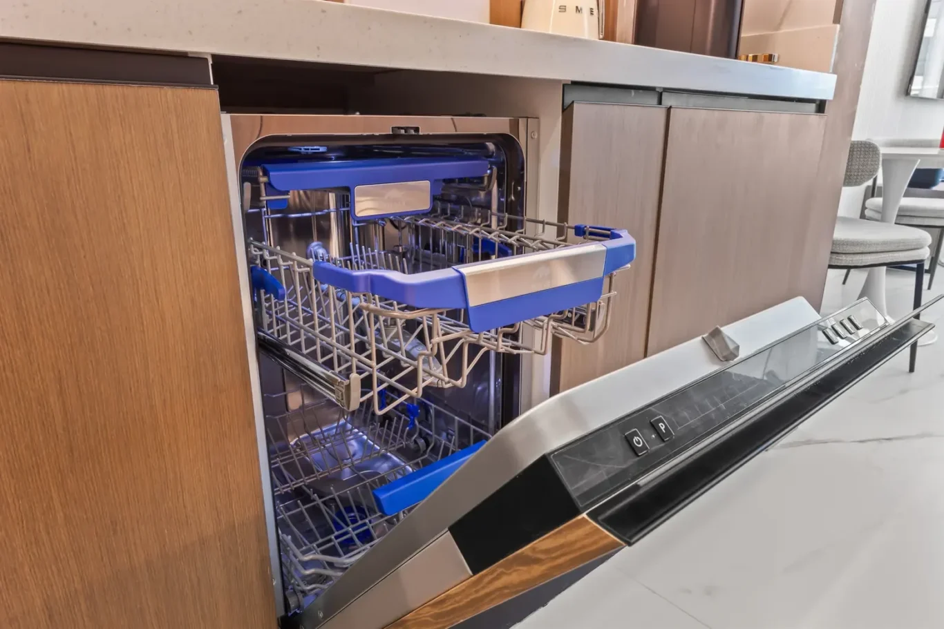 Open dishwasher with blue-rimmed rack pulled out, showing metal interior in a modern kitchen showroom.