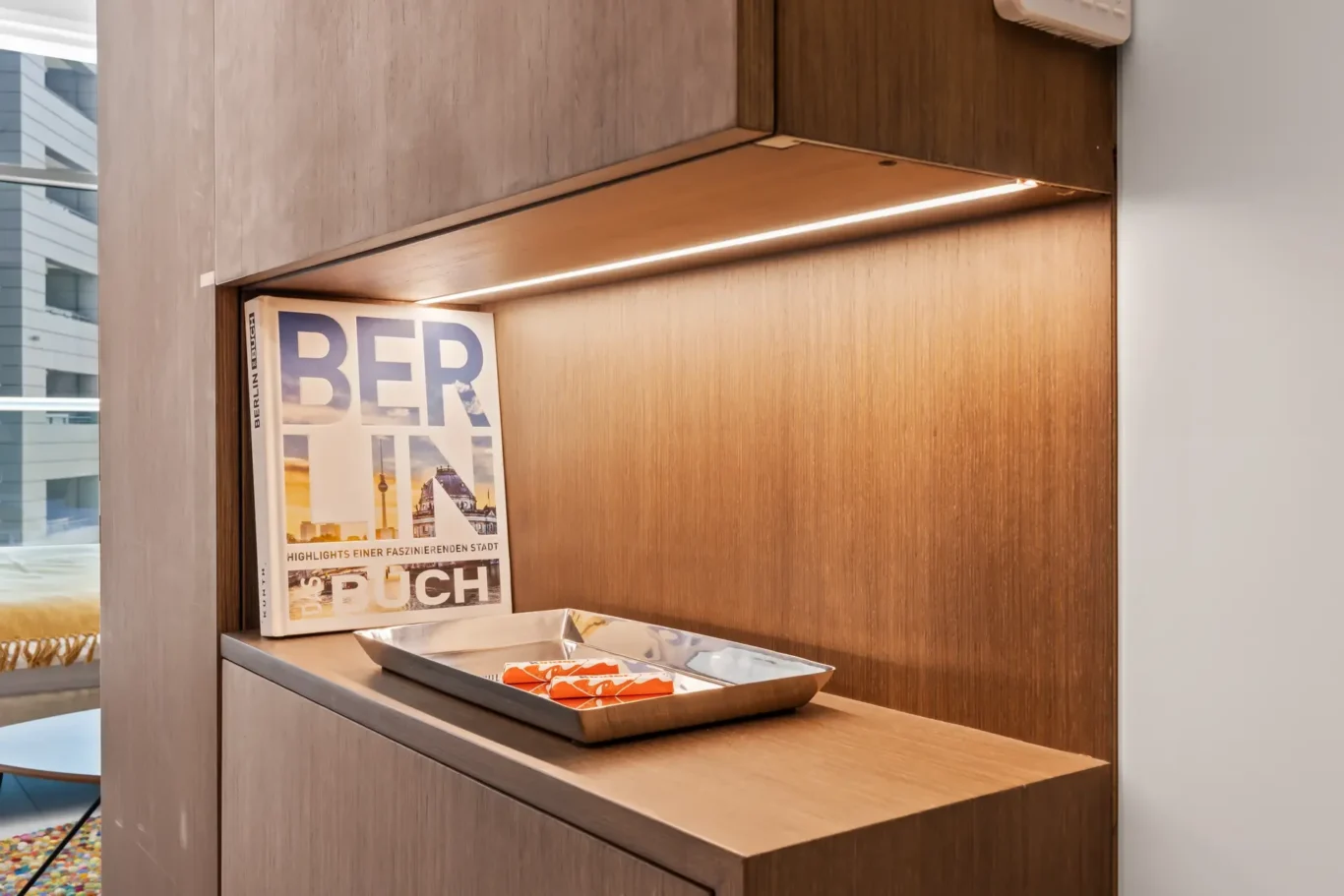 Wooden display shelf with a Berlin guide magazine in a lit recess and a metal tray of orange snacks beneath it.