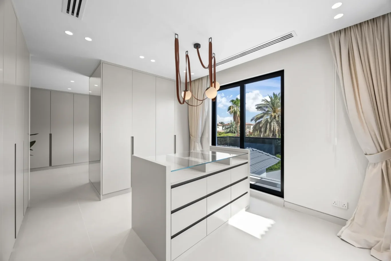 Modern white walk-in closet with tall cabinets, a glass-top island, and a large window showing palm trees outside.