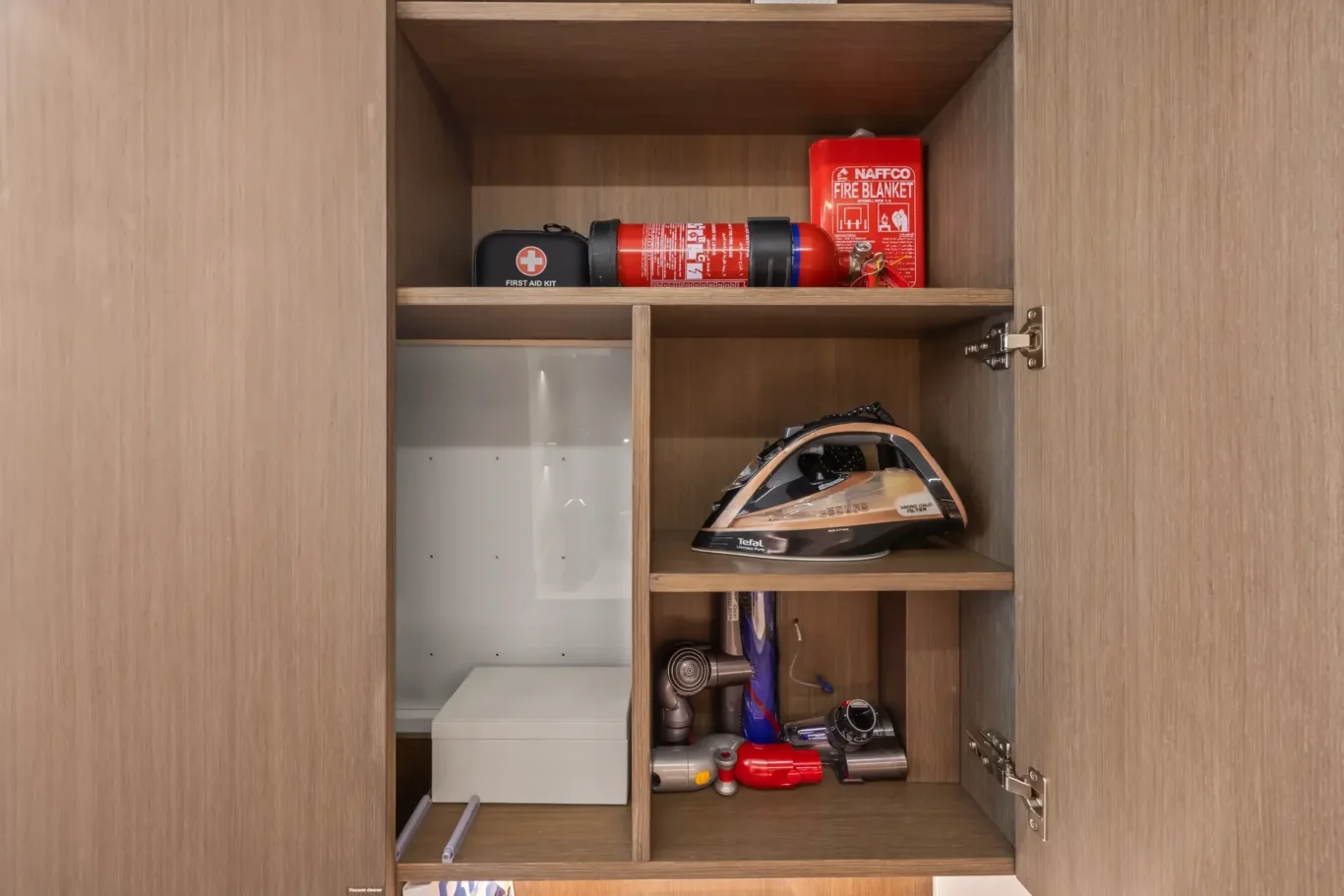 Open wooden cabinet showing a red fire blanket, a first aid kit, and other items on the top shelf.