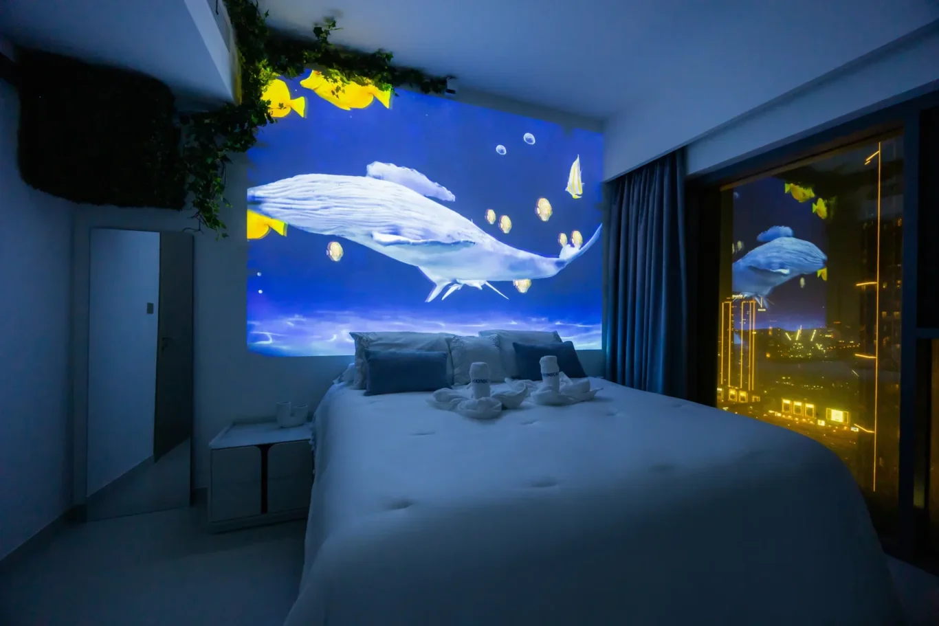 Hotel room at night with a large underwater mural of a whale shark and yellow fish behind the bed; city lights visible through a window on the right; white bedding and pillows.