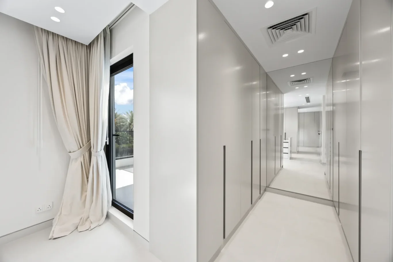 Bright modern hallway with floor-to-ceiling white wardrobes and a mirrored wall reflecting light and curtains edge paneling across a windowed door