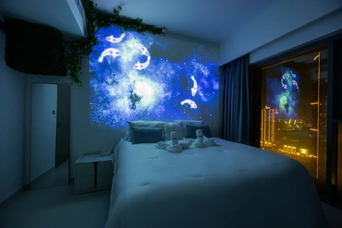 Hotel bedroom with a blue galaxy projection on the wall, bed neatly made with pillows and towels folded into swans; city lights visible through the window.