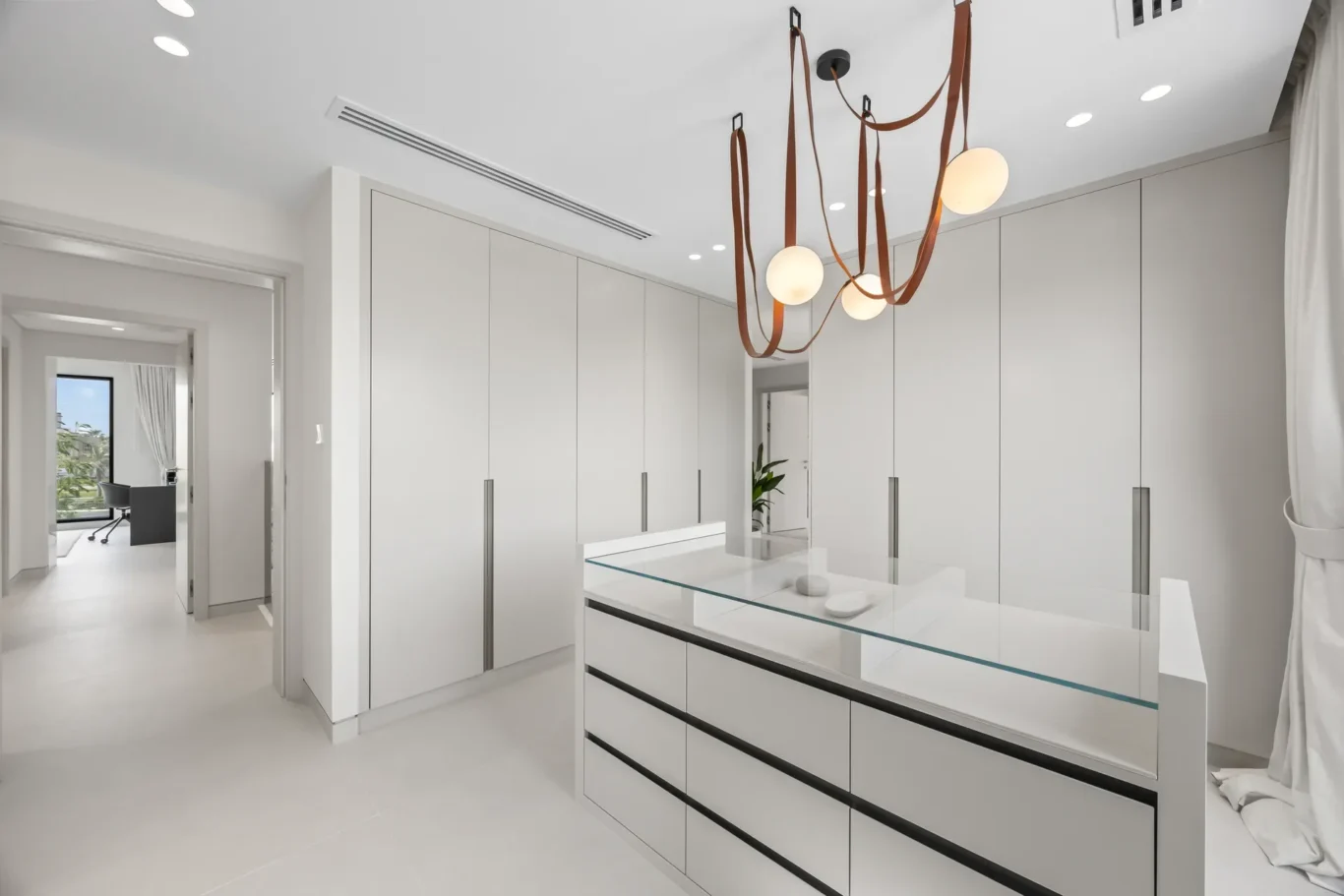 Modern white kitchen with tall cabinets and a glass-topped island; a sculptural leather-strapped pendant light hangs from the ceiling.