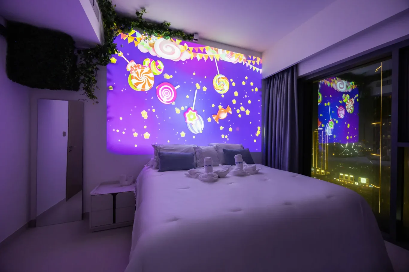 Hotel bedroom bathed in purple light with a candy-themed wall mural, plants along the ceiling, and a large bed with folded towel sculptures and cushions.