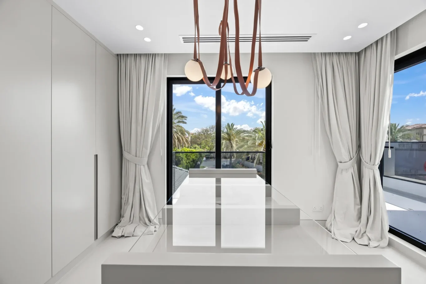 Bright minimalist dining area with a glossy white floor, white cabinetry, and a brown strap chandelier over a glass table facing a window view of palm trees.