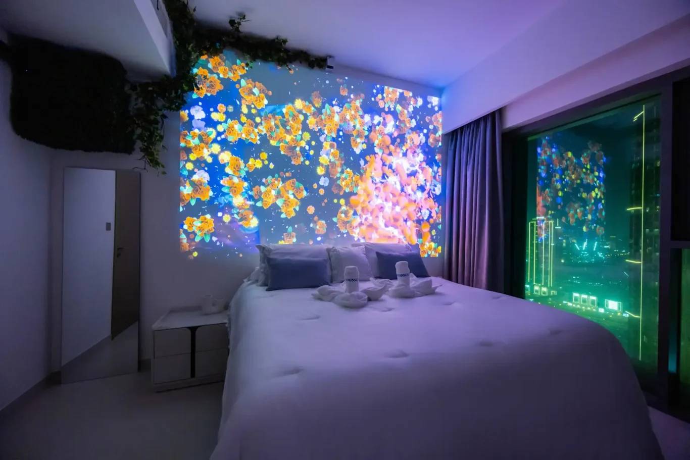 Hotel bedroom with a wall of orange flower projection above the bed and city lights outside the window.