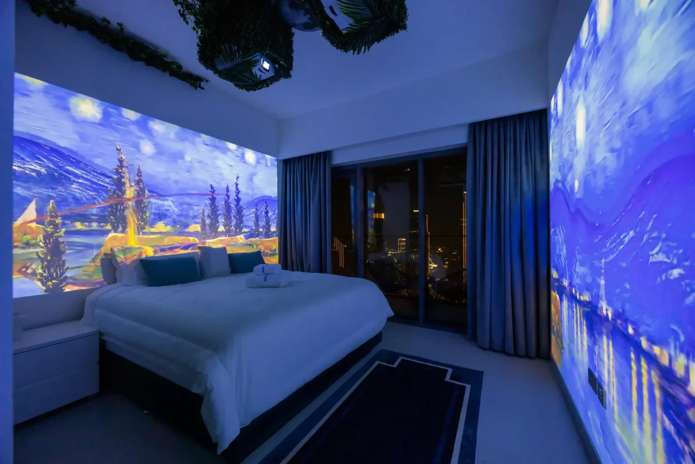 Hotel bedroom with a white bed and blue landscape projection on the walls, large windows, and city lights outside.