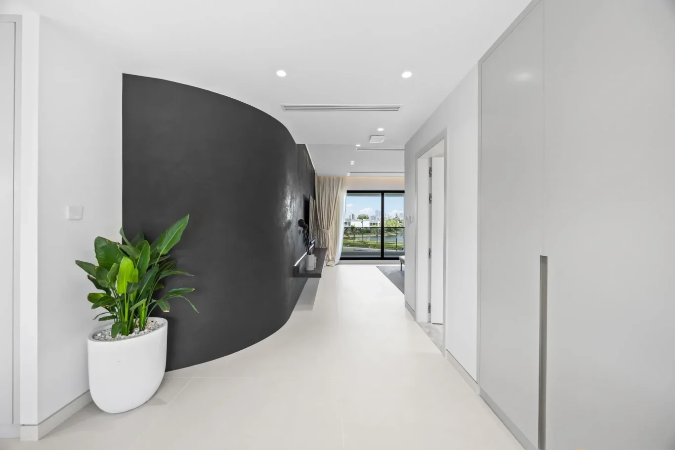 Modern hallway with a curved dark accent wall, white floor, and a large window showing a city view; potted plant in foreground left.