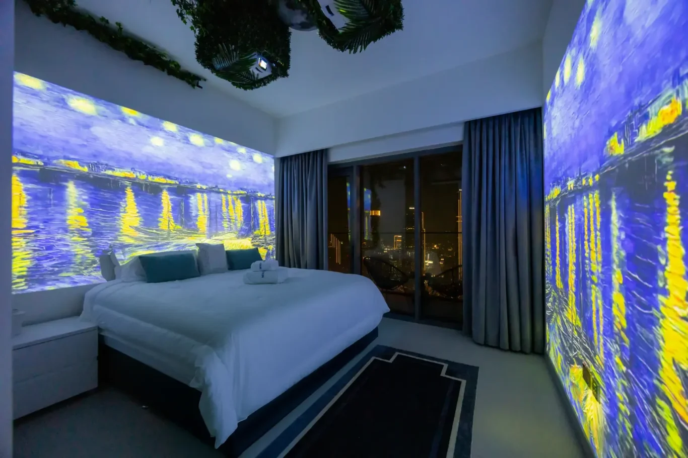 Modern bedroom with a large white bed, blue accent lighting on walls, and yellow highlights projected across the walls; glass doors open to a city skyline at night with dark curtains pulled to the sides.