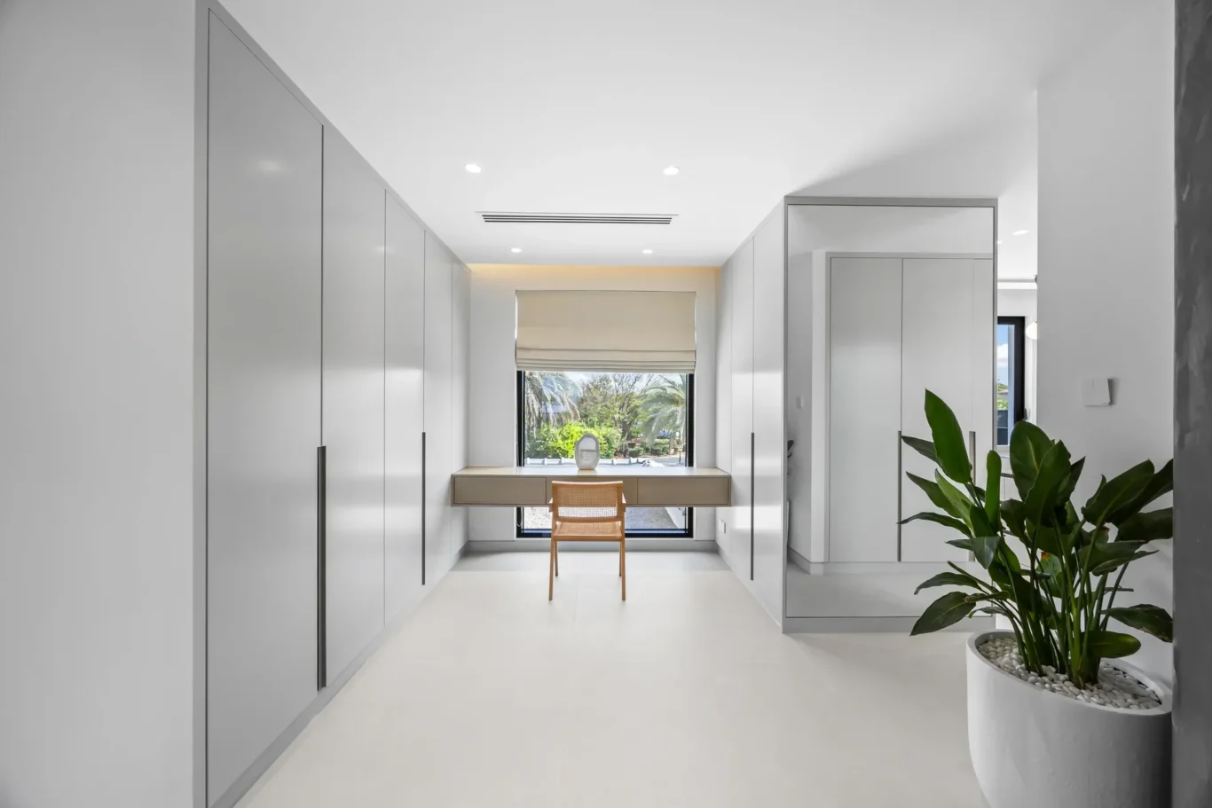 Bright white walk-in closet or dressing room with built-in wardrobes along both walls, a desk at the far end, and a large potted plant to the right. Window view shows greenery outside.