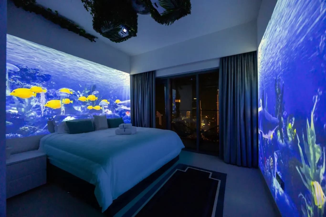 Bedroom with illuminated aquarium mural of yellow fish behind the bed, blue-tinted walls, and dark curtains open to a city skyline at night.