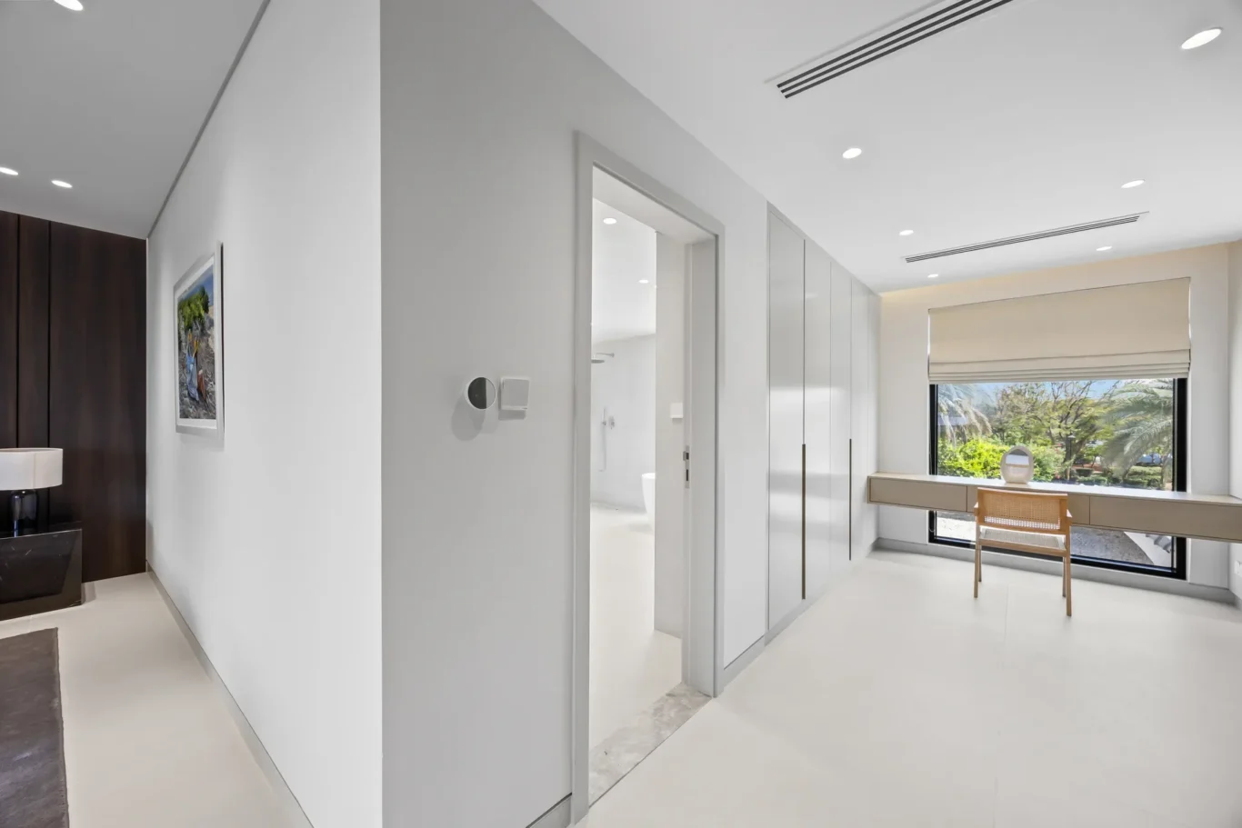 Bright, minimalist hallway with white walls and tall built-in white wardrobes along the right, and a desk by a large window showing greenery outside.