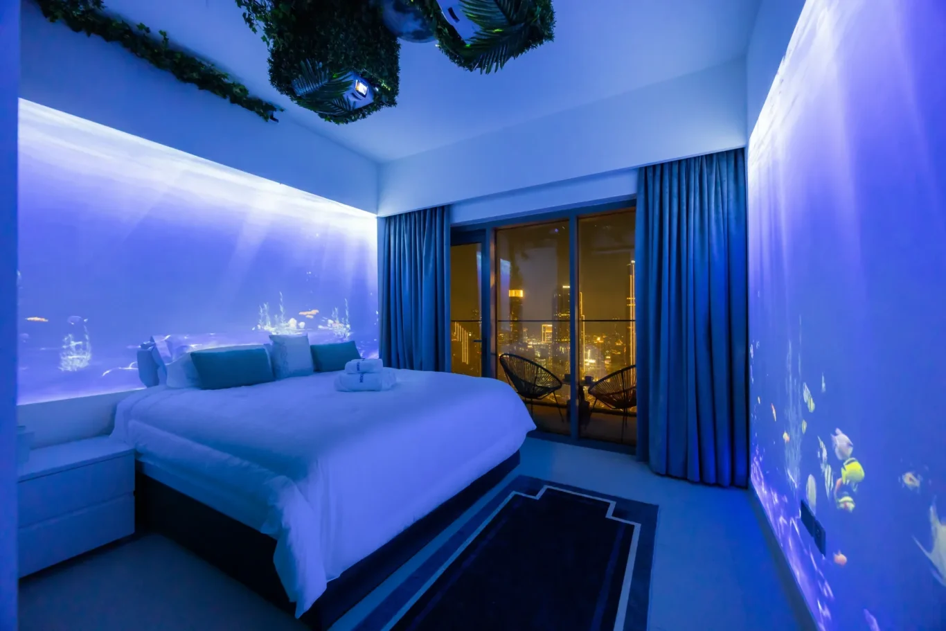 Blue-lit hotel room with a king-size bed, white linens and teal pillows, and city lights visible through glass doors.
