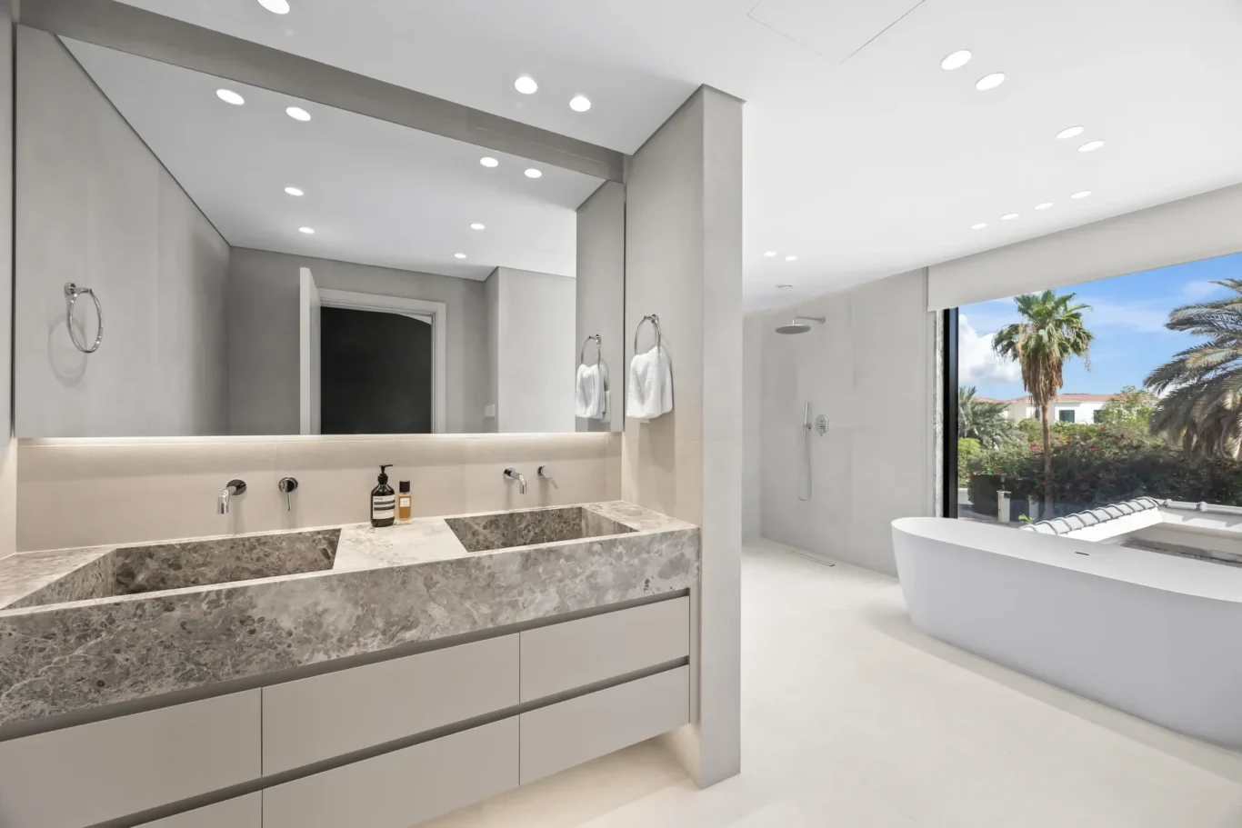Modern grey bathroom with a marble double vanity, large mirror, walk-in shower, and freestanding tub by a window