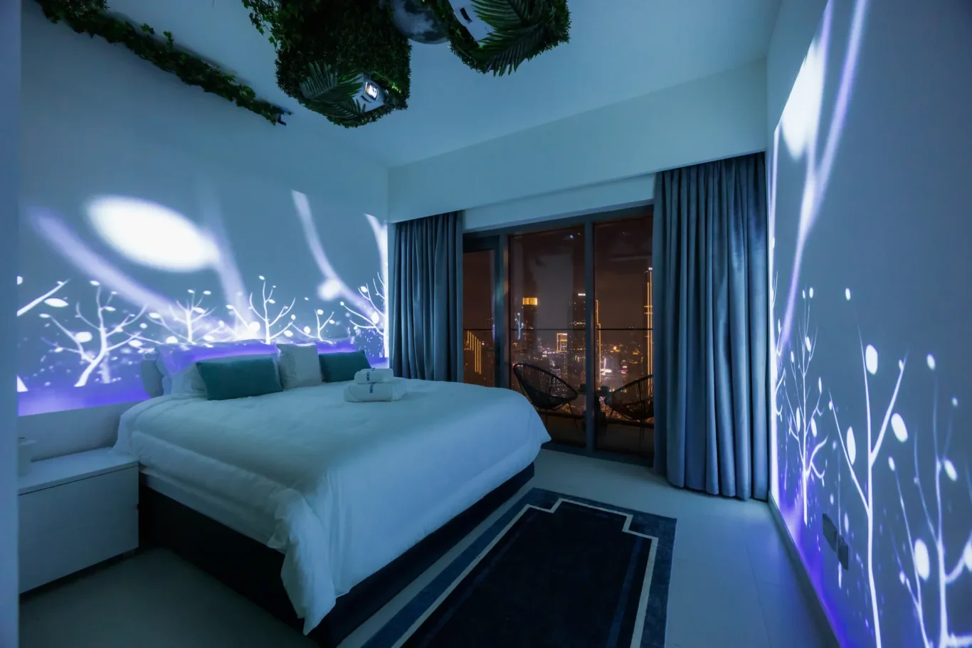 King-size bed in a modern hotel room with white bedding, teal pillows, and blue light projections of tree branches on the walls; city lights visible through glass doors.