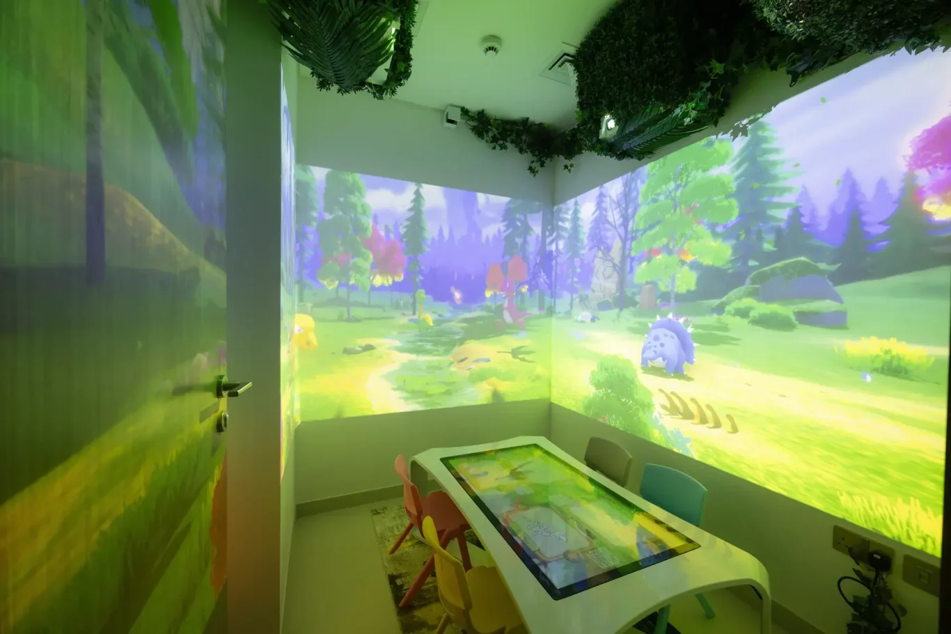 Children's playroom with a forest projection on the walls and a touchscreen table surrounded by small chairs.