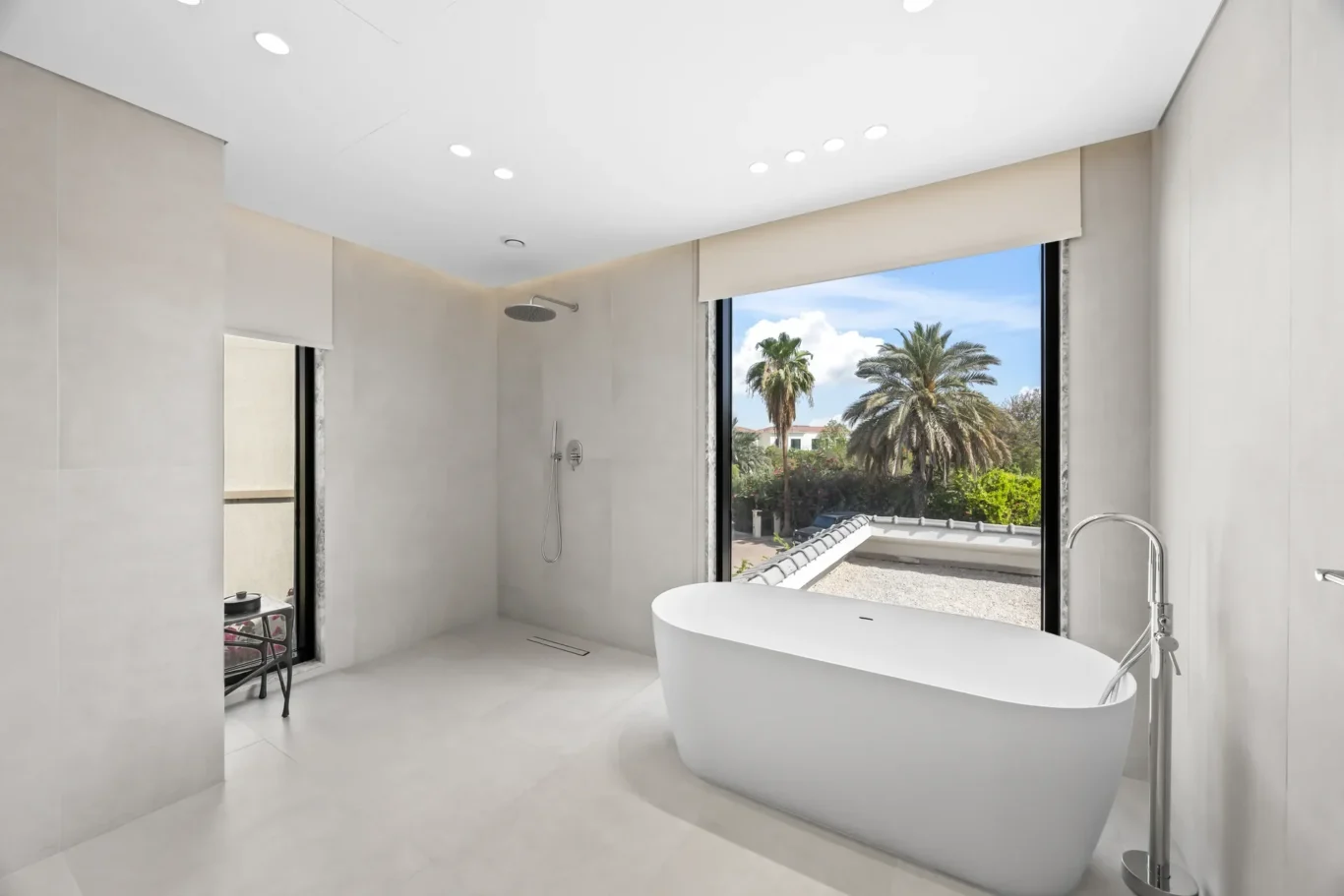 Luxurious bathroom with a white freestanding tub, glass shower, and a large window overlooking palm trees outside.