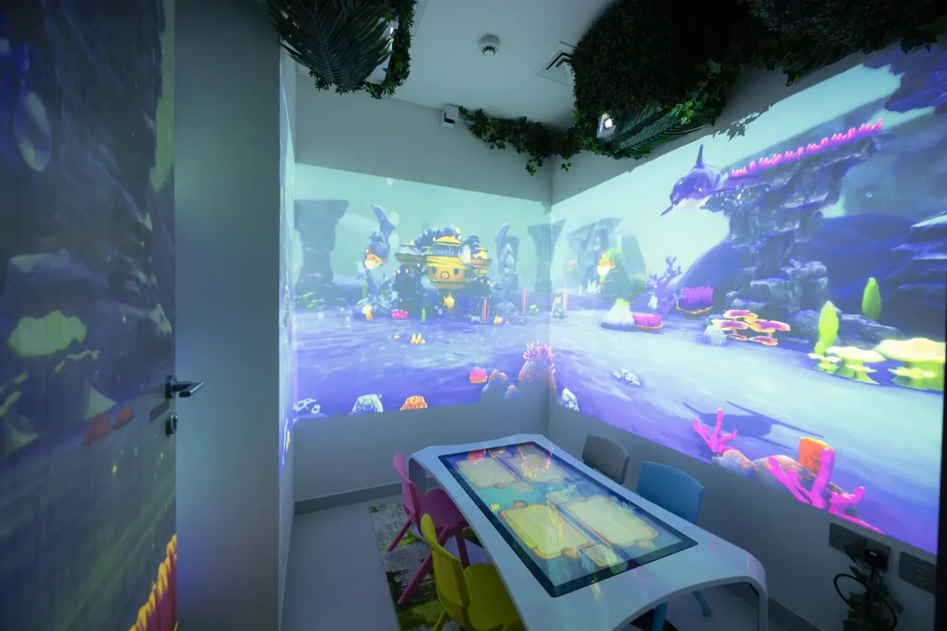 Room with an underwater projection mural across the walls, a touch-screen game table, and colorful child-sized chairs around it.