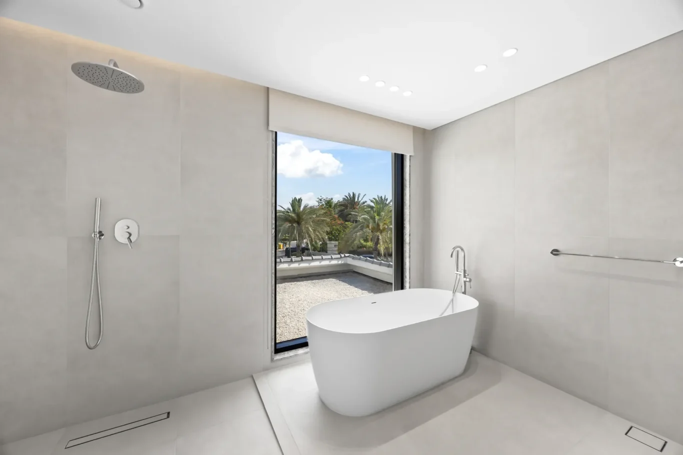 Modern bathroom with a freestanding white tub beside a large window showing palm trees outside; rainfall showerhead and chrome fixtures above a light-gray tiled wall.