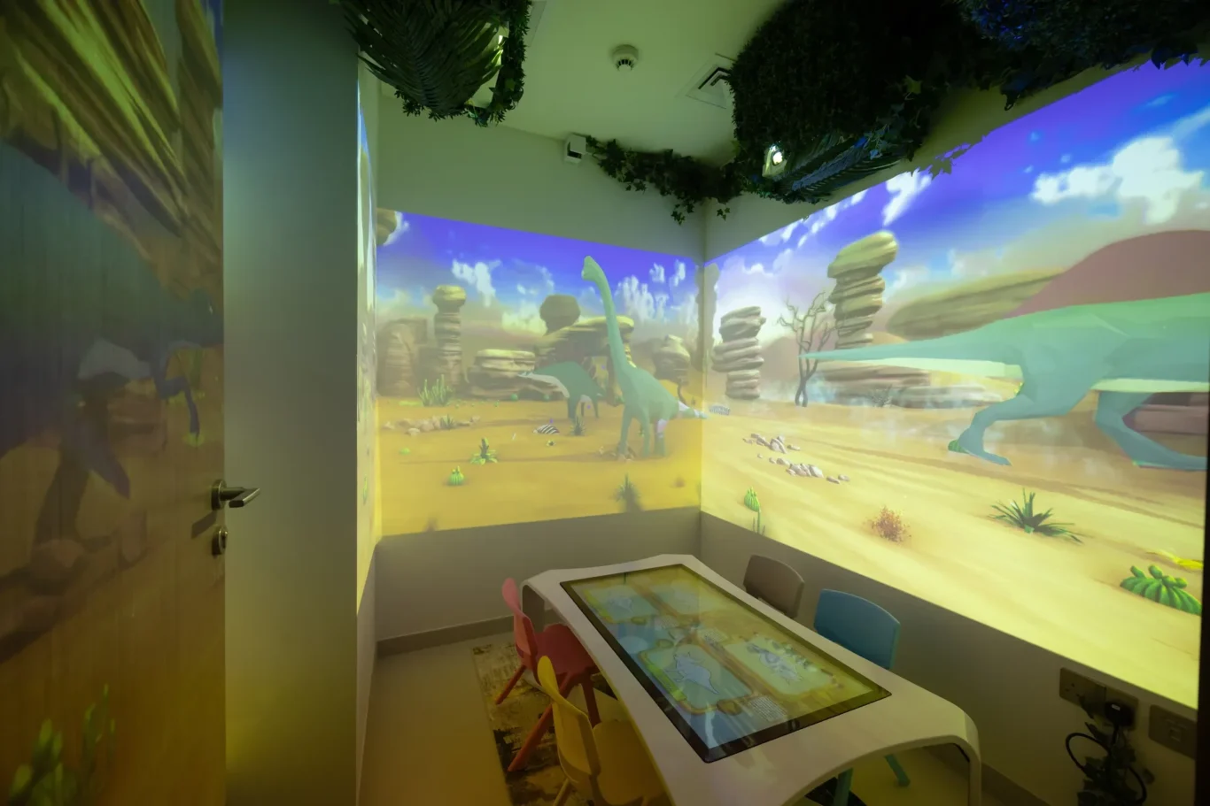Colorful kids' playroom with a dinosaur desert mural, a white interactive table, and small chairs.