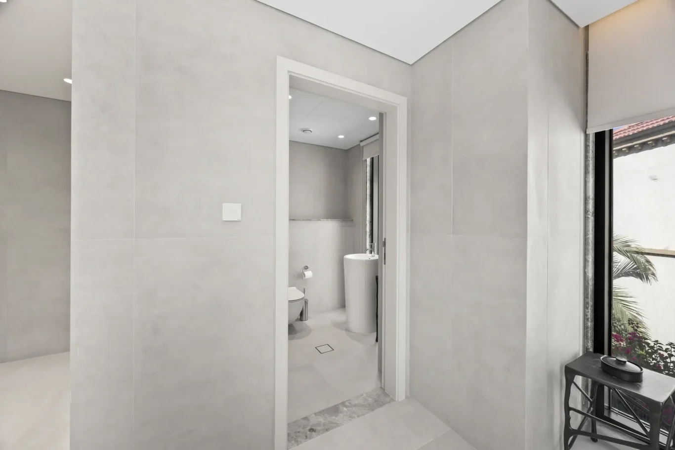 Modern gray-tiled bathroom seen through an open doorway, featuring a toilet, pedestal sink, and recessed ceiling lights.