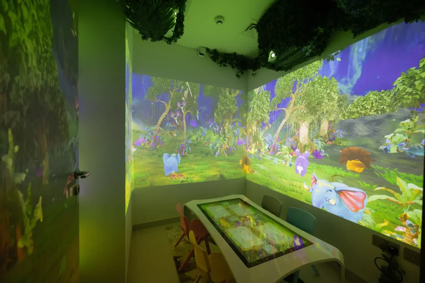 Playroom with immersive forest projection on walls and ceiling, featuring colorful animals and a white touch-table with chairs nearby.