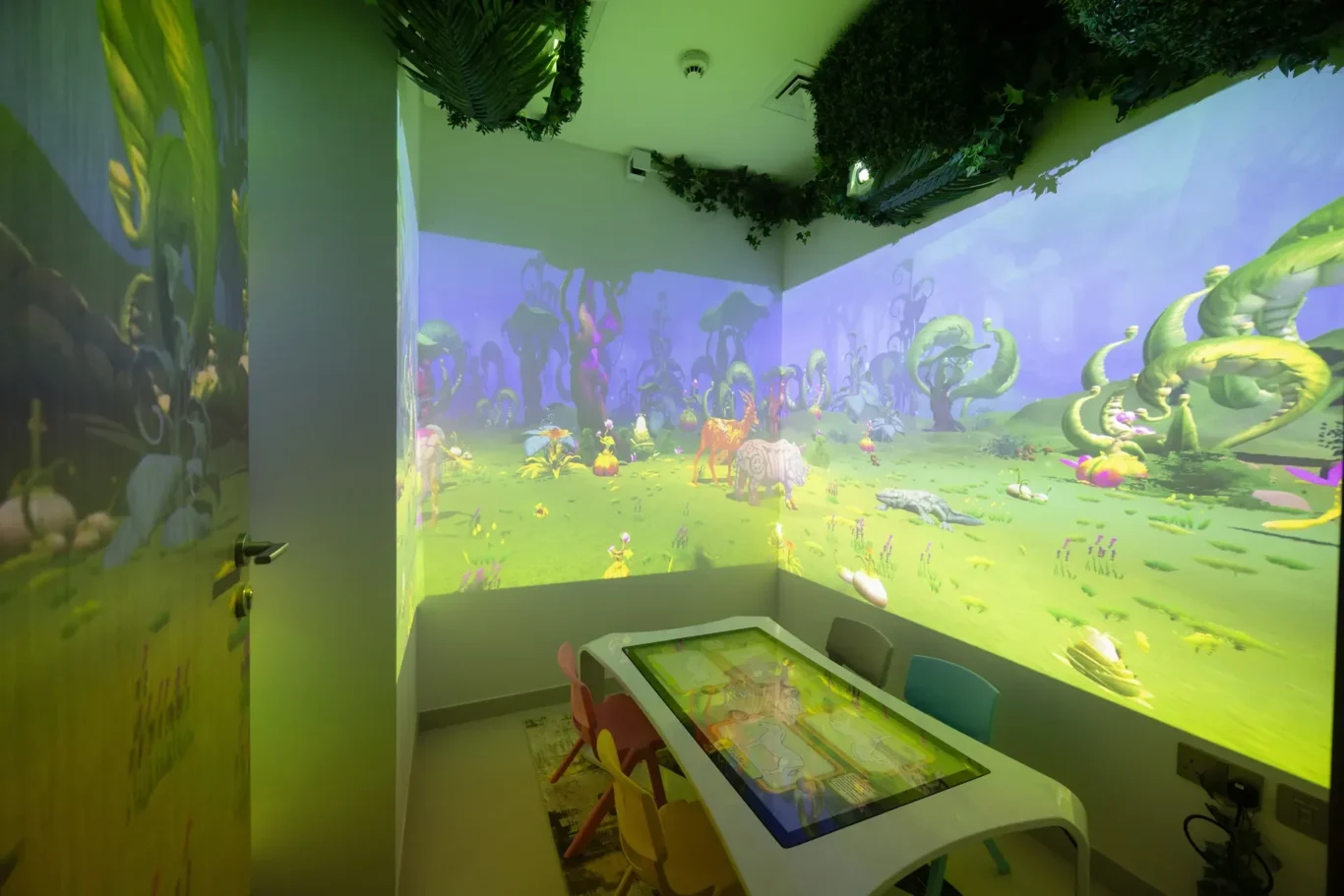 Small playroom with a bright jungle mural projected on the walls, ceiling plants, and a white interactive touchscreen table with colorful chairs.