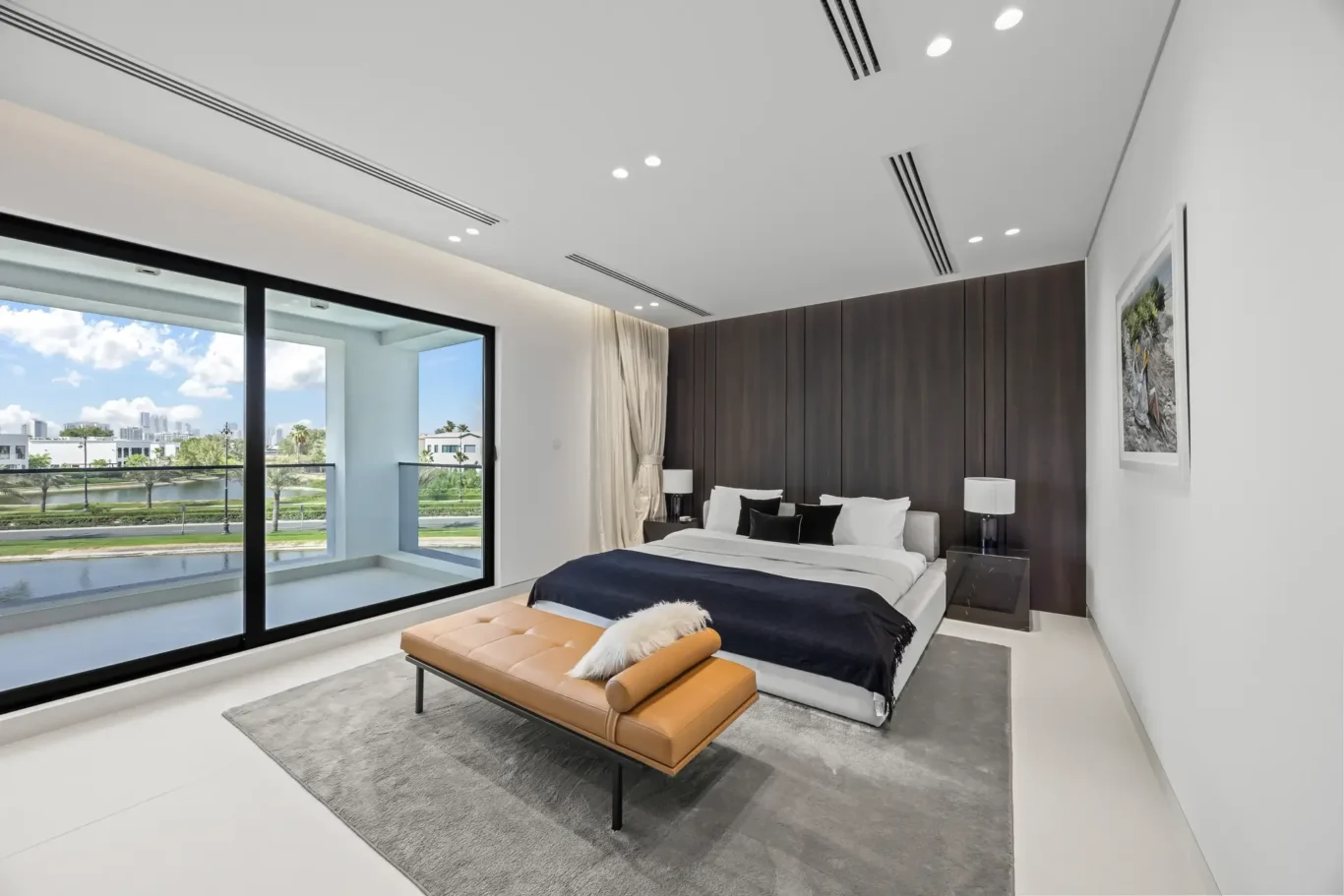 Modern bedroom with a large glass wall showing a city view, a bed with dark bedding, and a tan bench at the foot.