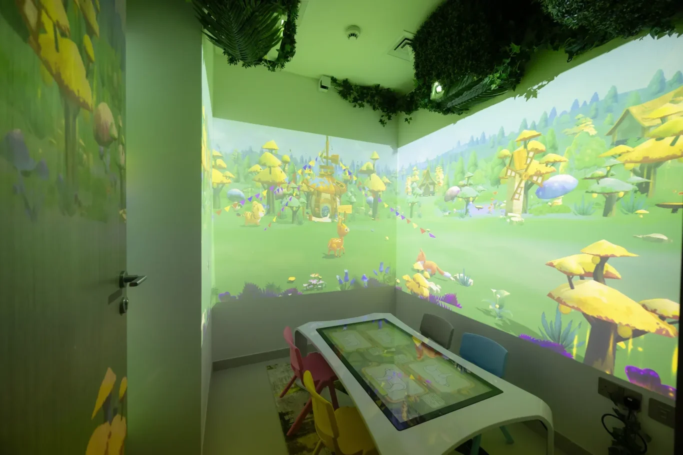 Playroom with a green forest mural wrapping around walls and a white interactive touch table for kids.