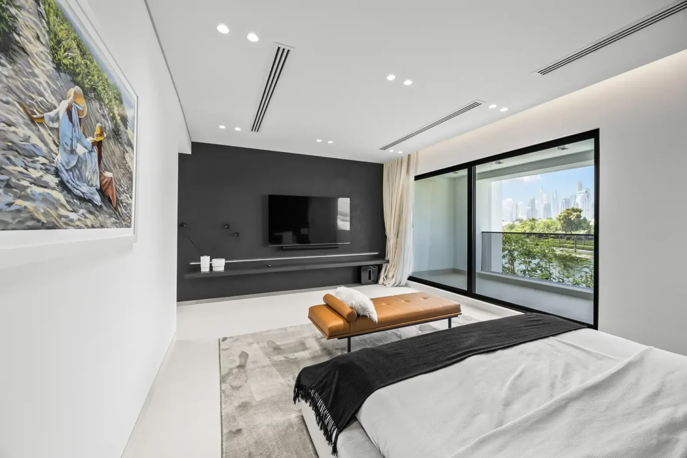 Modern bedroom with a wall-mounted TV on a dark accent wall, a tan bench, and a balcony view through sliding doors of the room's right side.