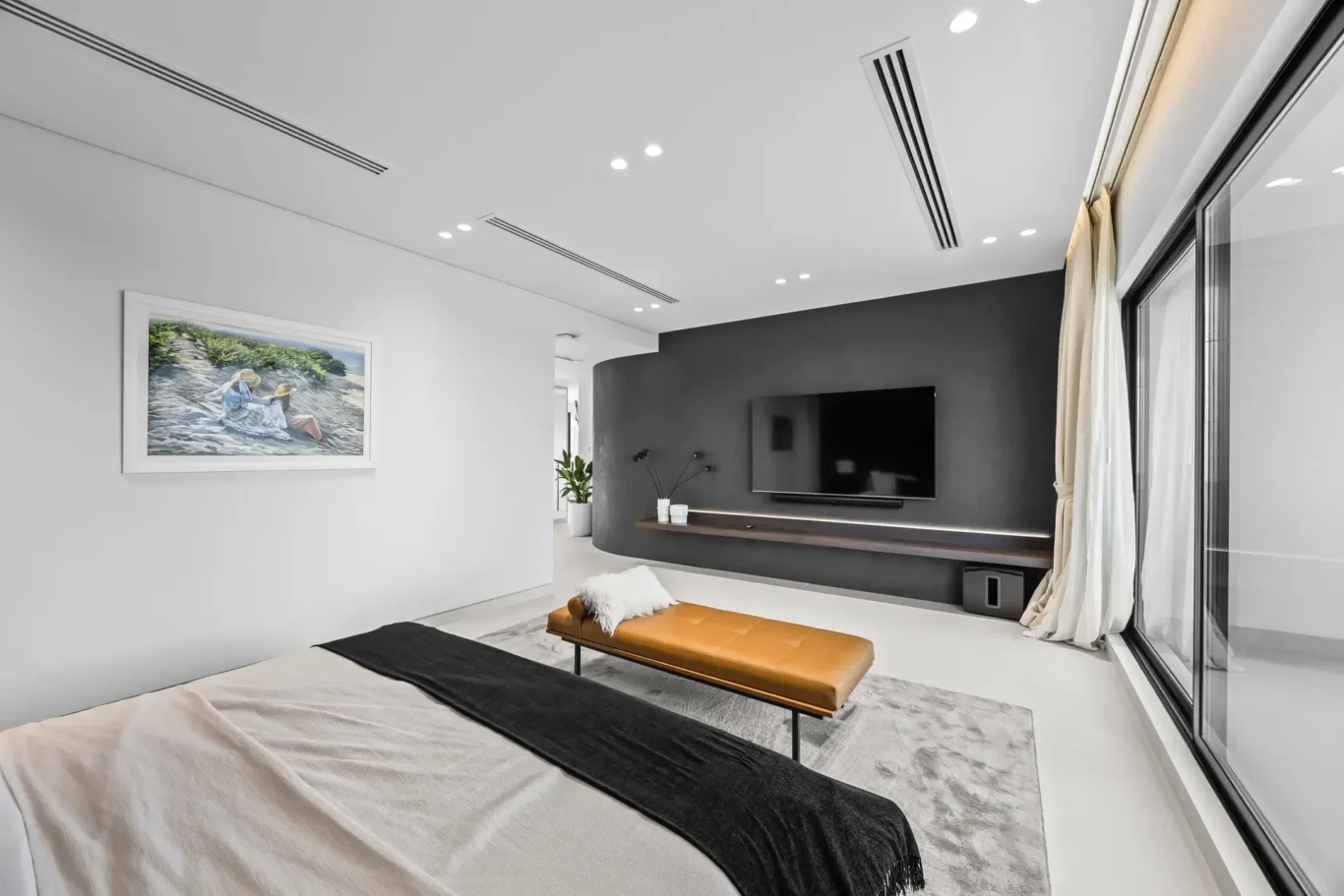 Contemporary bedroom and lounge area with a dark wall-mounted TV, orange leather bench, white walls, and large glass doors with curtains on the right.