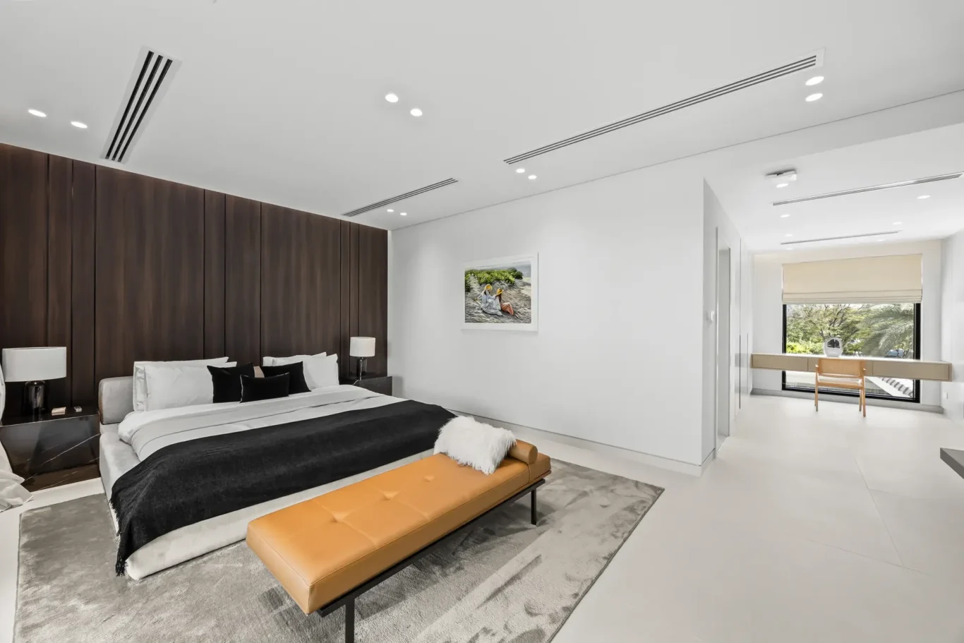 Modern hotel bedroom with a large bed, dark wooden headboard, white and black bedding, and a tan bench at the foot of the bed