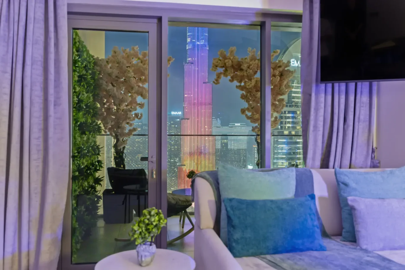 Living room with glass doors showcasing a night-city skyline, purple lighting, and a sofa with blue cushions.