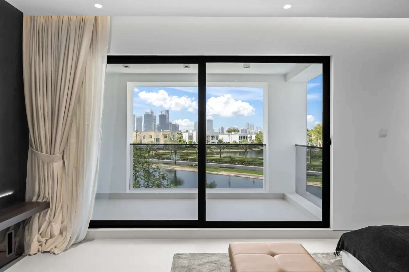 Modern bedroom with large black-framed sliding glass doors opening to a balcony and cityscape beyond, curtains pushed to the side, neutral decor, and a white floor.
