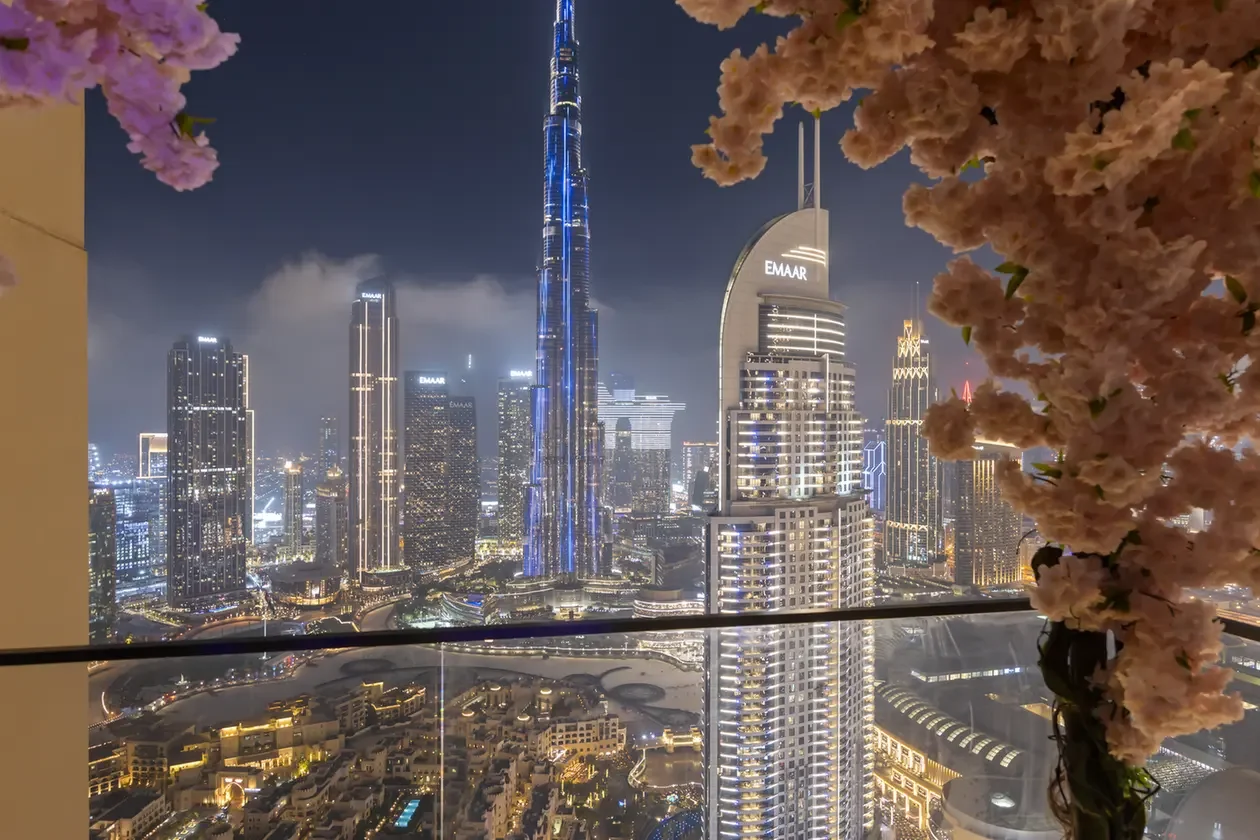 Nighttime Dubai skyline with illuminated skyscrapers, seen from a balcony framed by pink blossoms and a glass railing.