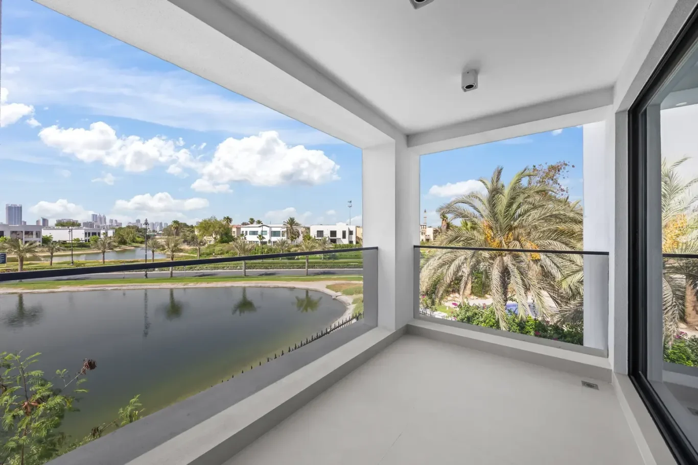 Balcony with white walls and glass railing overlooking a pond, palm trees, and distant city skyline under a blue sky.