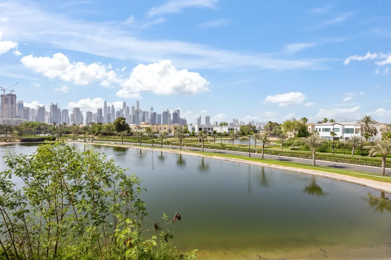 Urban waterfront park with a calm canal in the foreground and modern skyscrapers in the distance under a blue sky.
