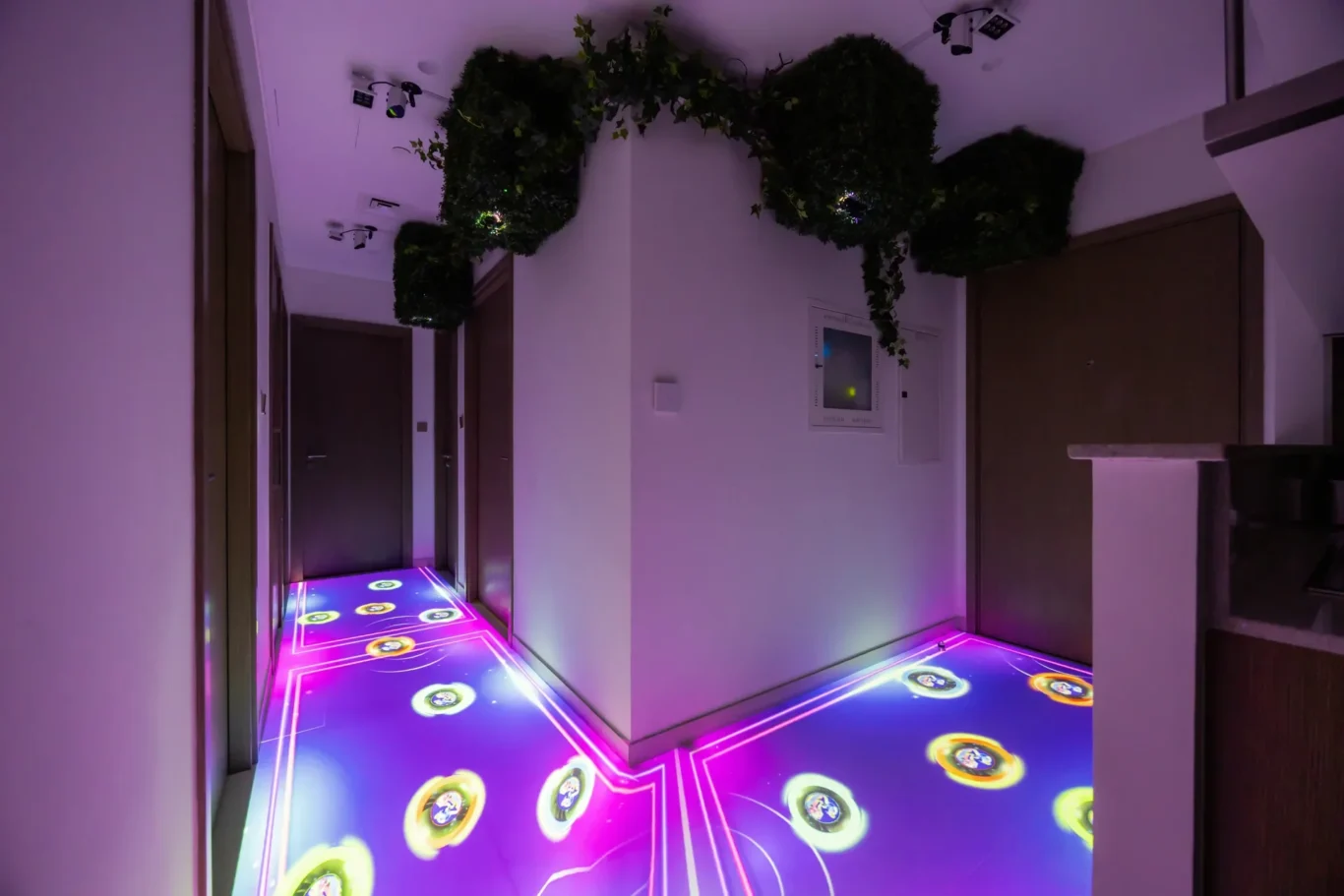 Indoor hallway with purple neon floor projection showing circular icons and glowing geometric lines, plants along the ceiling edge, and closed doors on both sides.