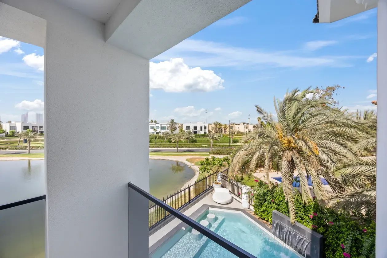 View from a balcony overlooking a rectangular pool, glass railing, palm trees, and a pond in a sunny residential development.