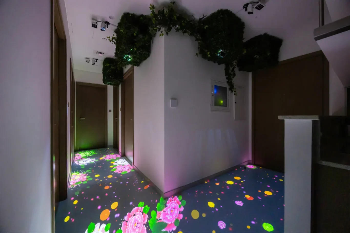 Hallway with hanging greenery and a colorful floral projection on the floor, plus doors along the corridor, in a dimly lit setting.
