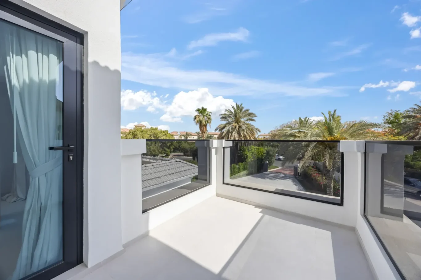 Balcony view with glass railing, white building, and a blue sky, palm trees beyond the railing, and a door with blue curtains on the left.